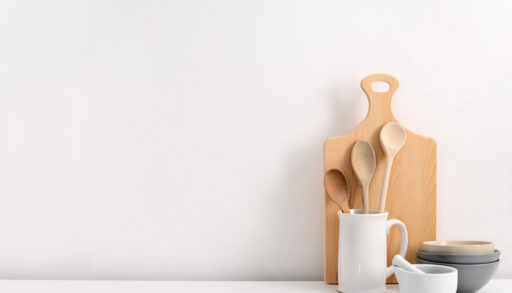 Wooden cutting board and white jar with wooden spoons on a white countertop.