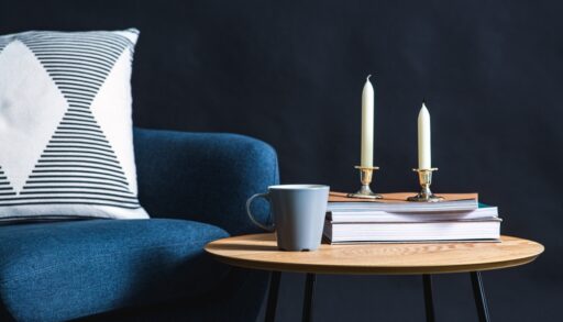 Blue armchair next to a wooden table with a stack of books, candles and a grey coffee cup.