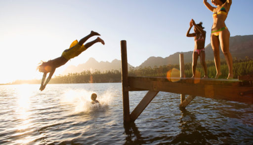 Profile shot of a father and son jumping off a dock into the lake at sunset with a splash as the daughter and mother cheers on, share a rental