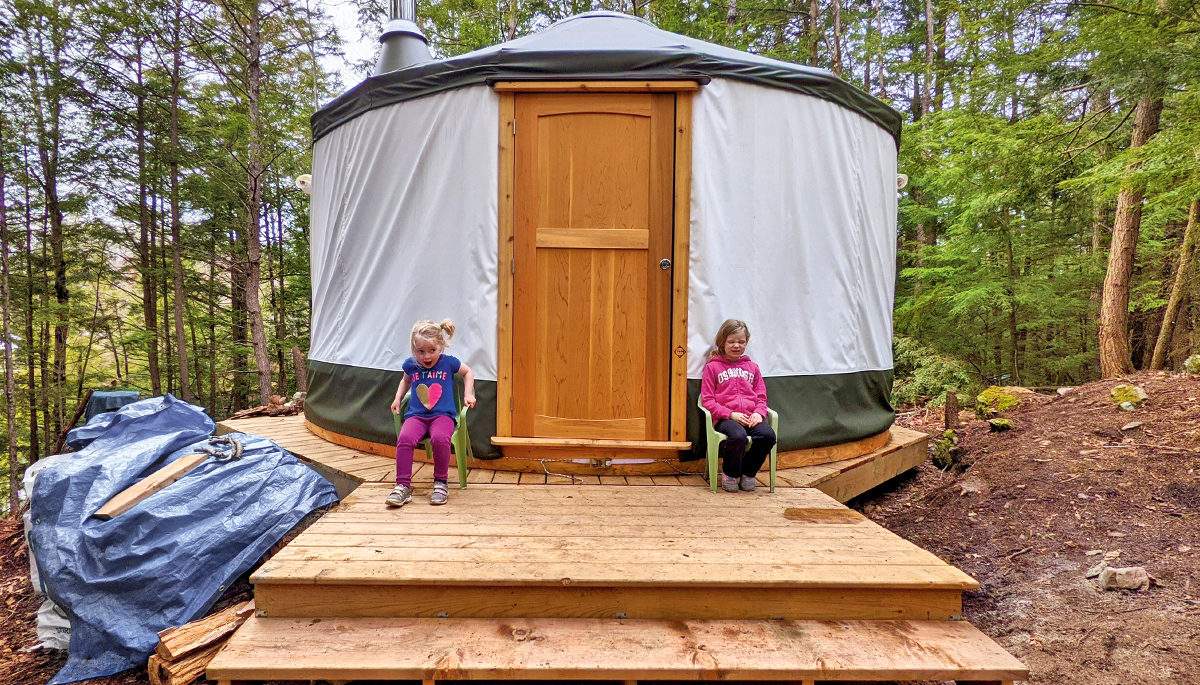 two small girls sitting outside of a yurt built on a wooden platform in a forest