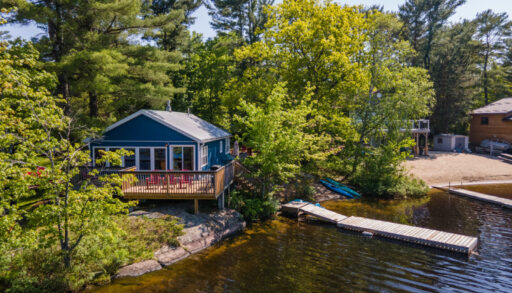 A small blue cottage with a big deck, a sandy shore, and a long dock sits lakeside. Green trees surround the land behind..