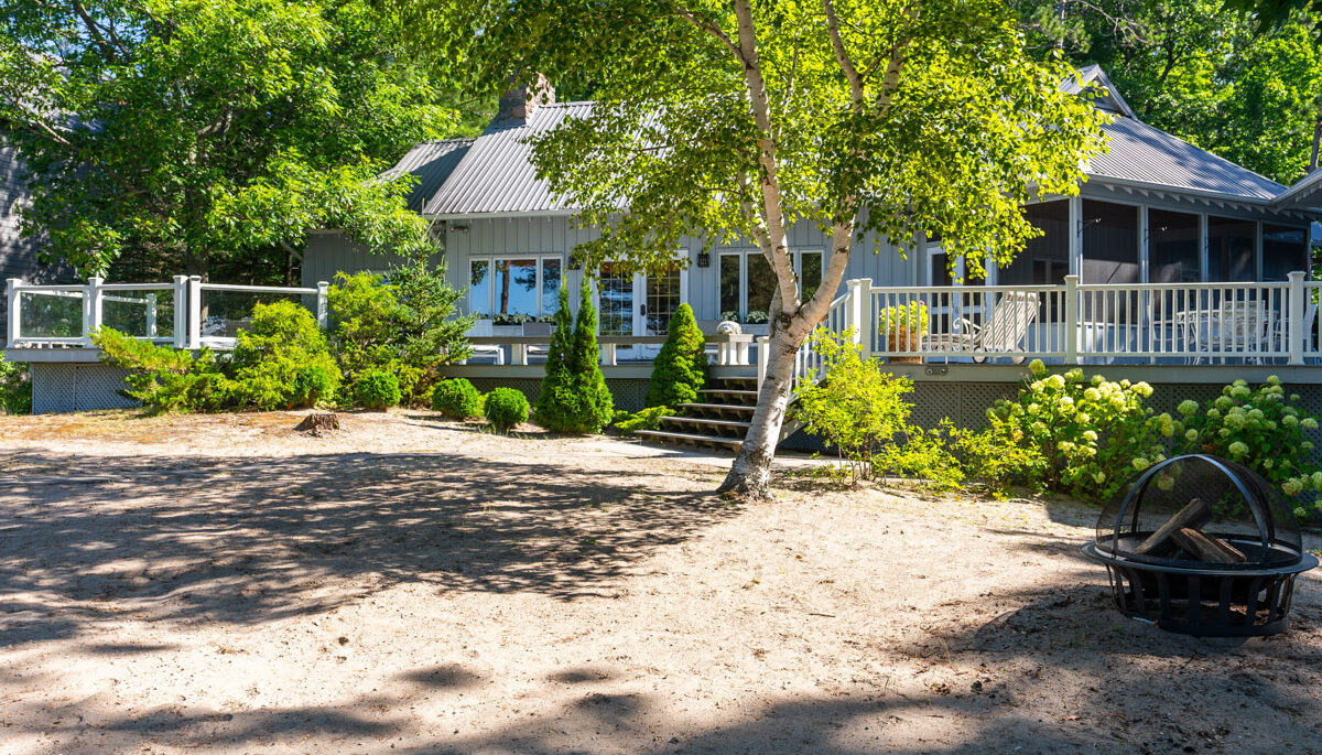 A wide, sandy area lined in greenery, in front of a large luxury cottage.