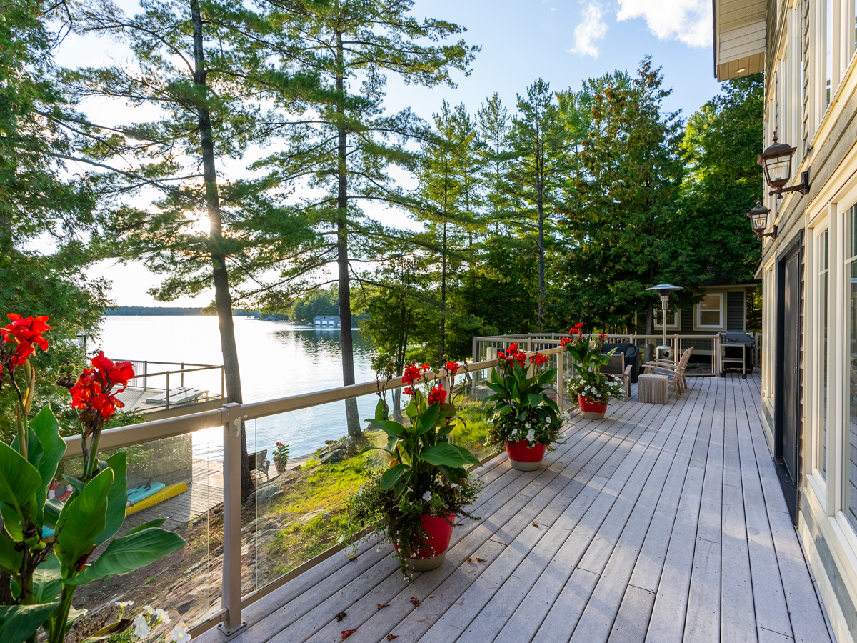 A deck with a glass railing and lake views, lined with potted flowers.