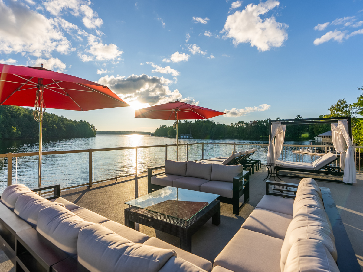 A large rooftop deck with outdoor living furniture and red sun umbrellas, looking out over a lake.