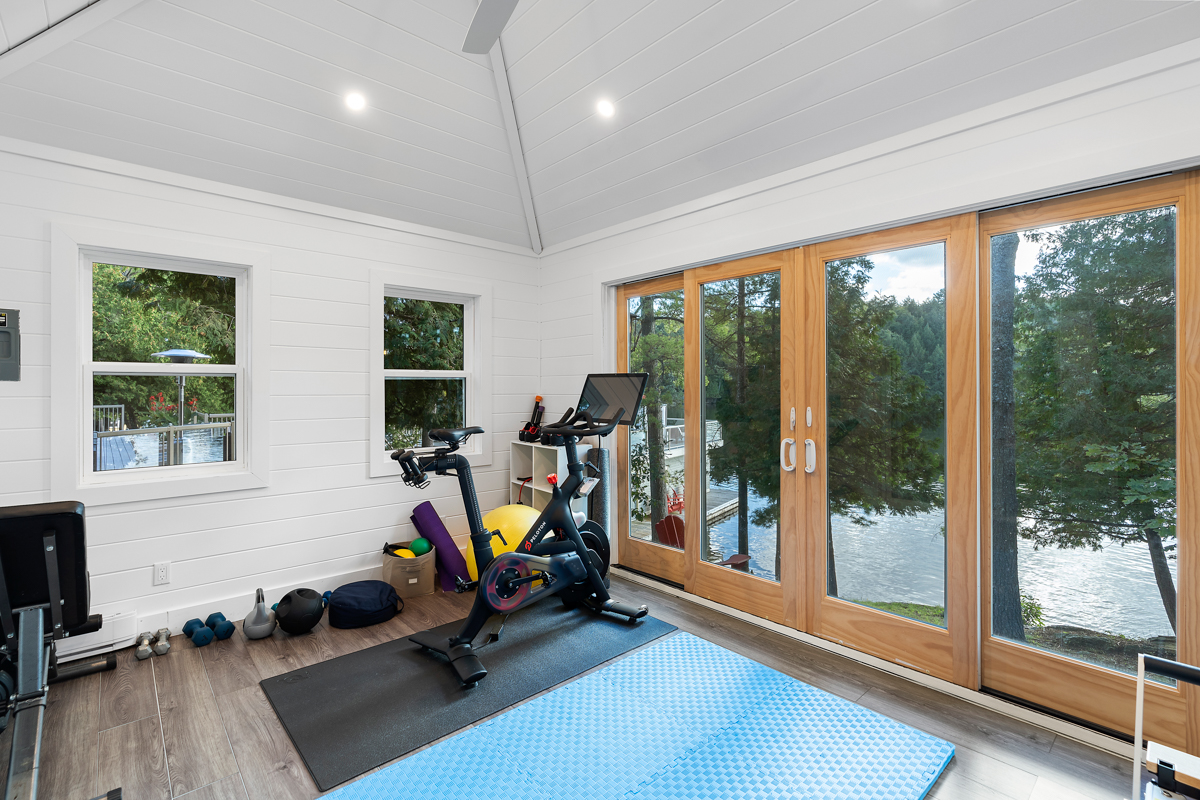 A detached home gym space with workout equipment and floor-to-ceiling windows that look out to a lake view.