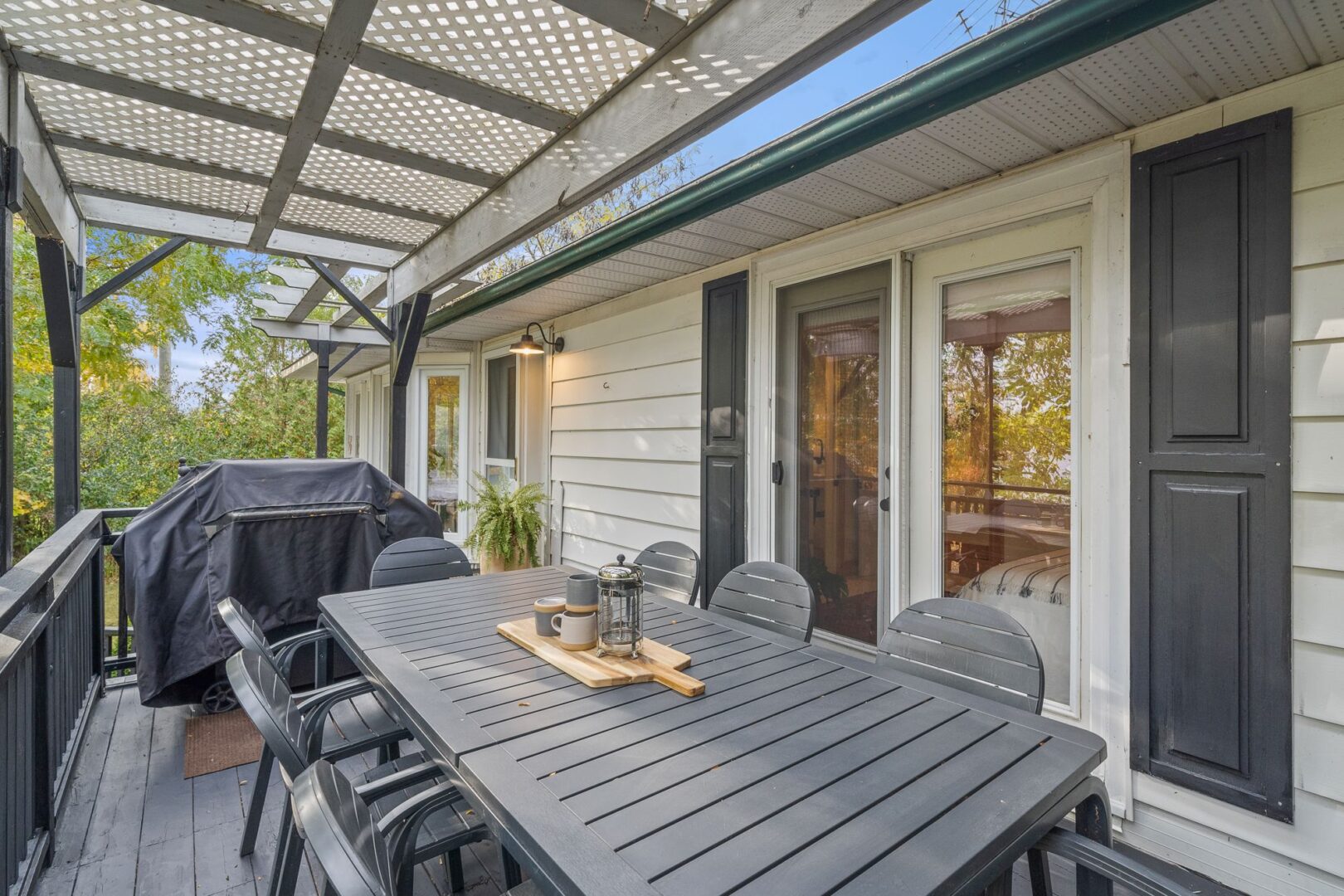 A barbecue and an outdoor dining table and chairs sit on the front porch of a raised bungalow.