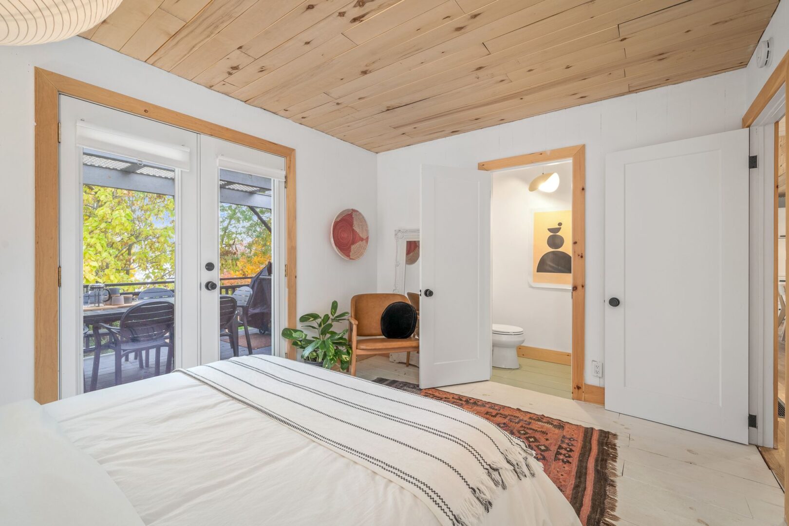 A big, bright bedroom with a large bed, a wood-panelled ceiling, a small ensuite bathroom, and double glass doors leading to a deck.