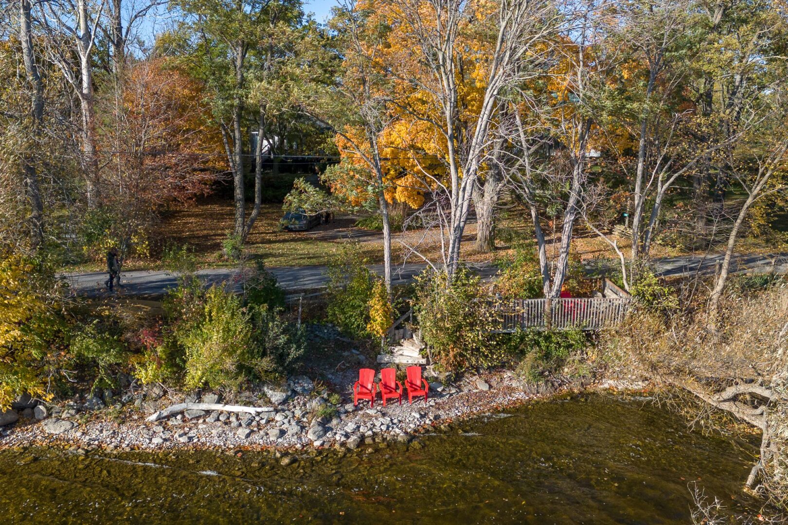 Two red Muskoka chairs sit on the rocky shore of a lake, just in front of a residential road.