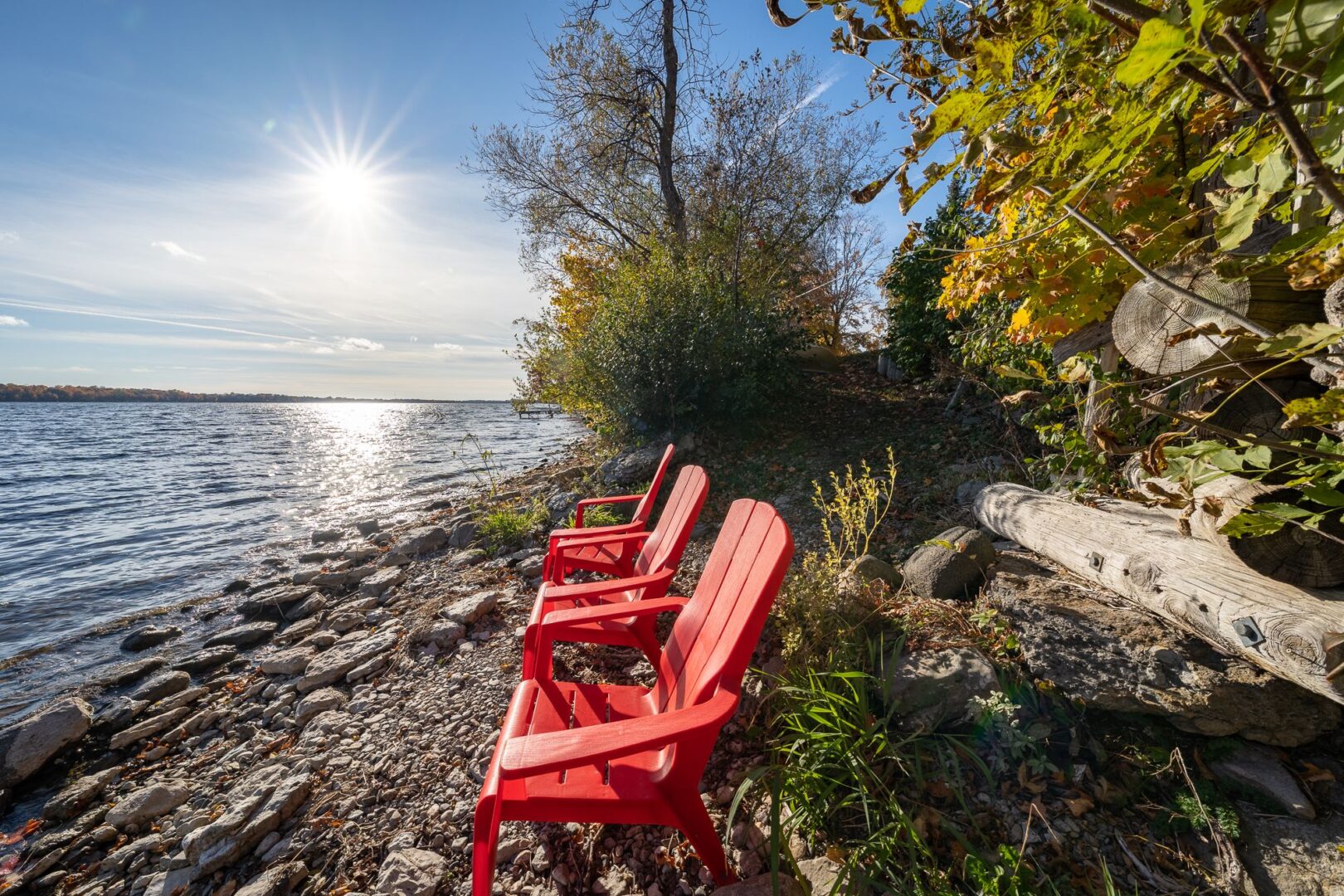Two red Muskoka chairs sit on the rocky shoreline of a sunny stretch of lake.