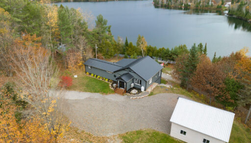 A small bungalow with a blue-gray roof and siding sits waterfront on a calm lake. The front of the house has a large paved driveway, and the home is surrounded by trees and greenery.