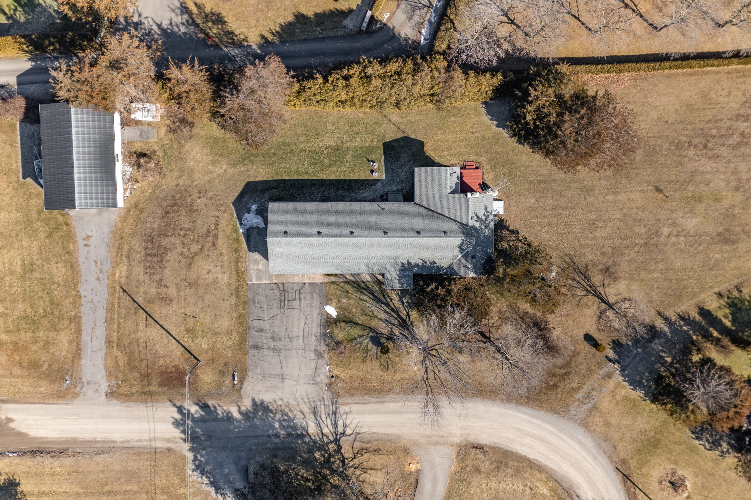 Aerial view of a bungalow with a gray roof, a long driveway, and a detached garage, sitting on a grassy lot.