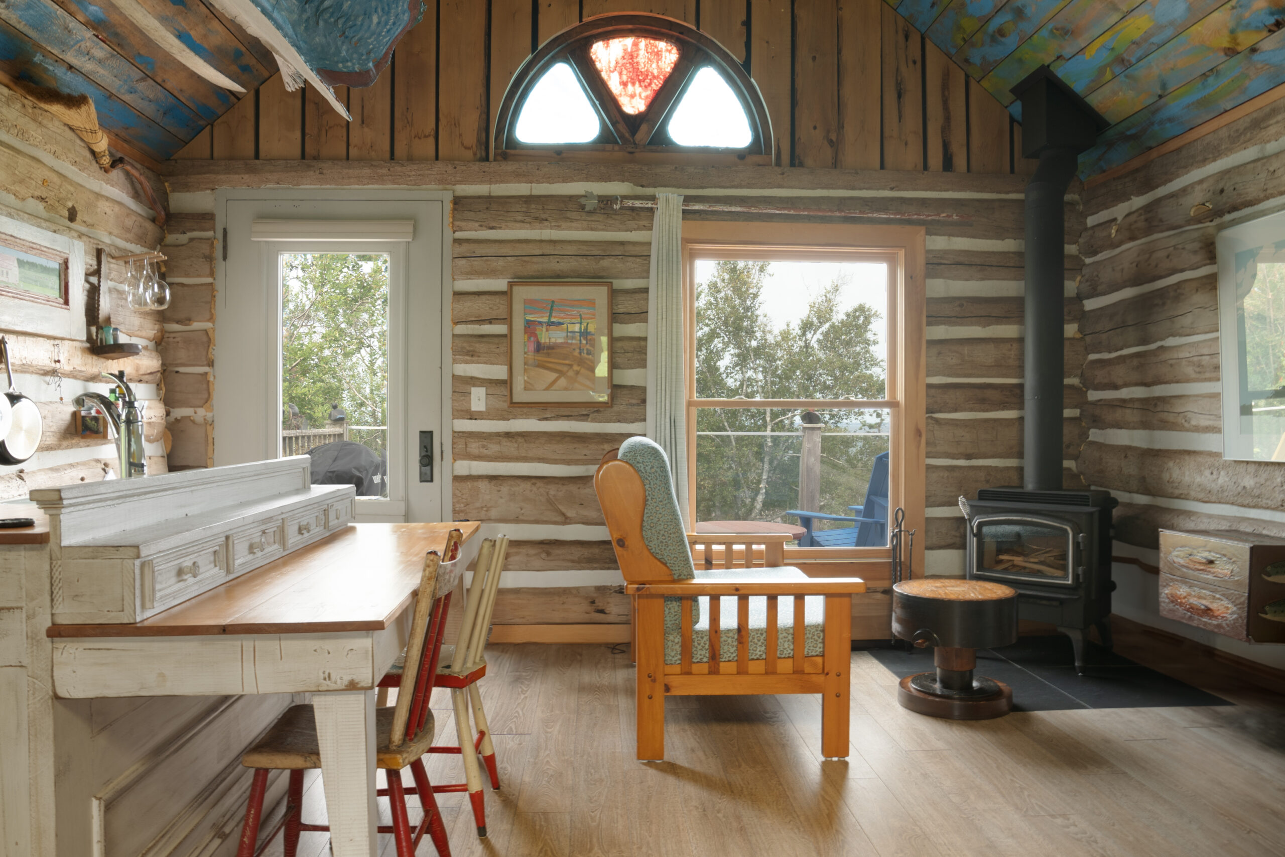 Interior of a small log cabin, with big windows, a small table, a wood stove, and a chair.