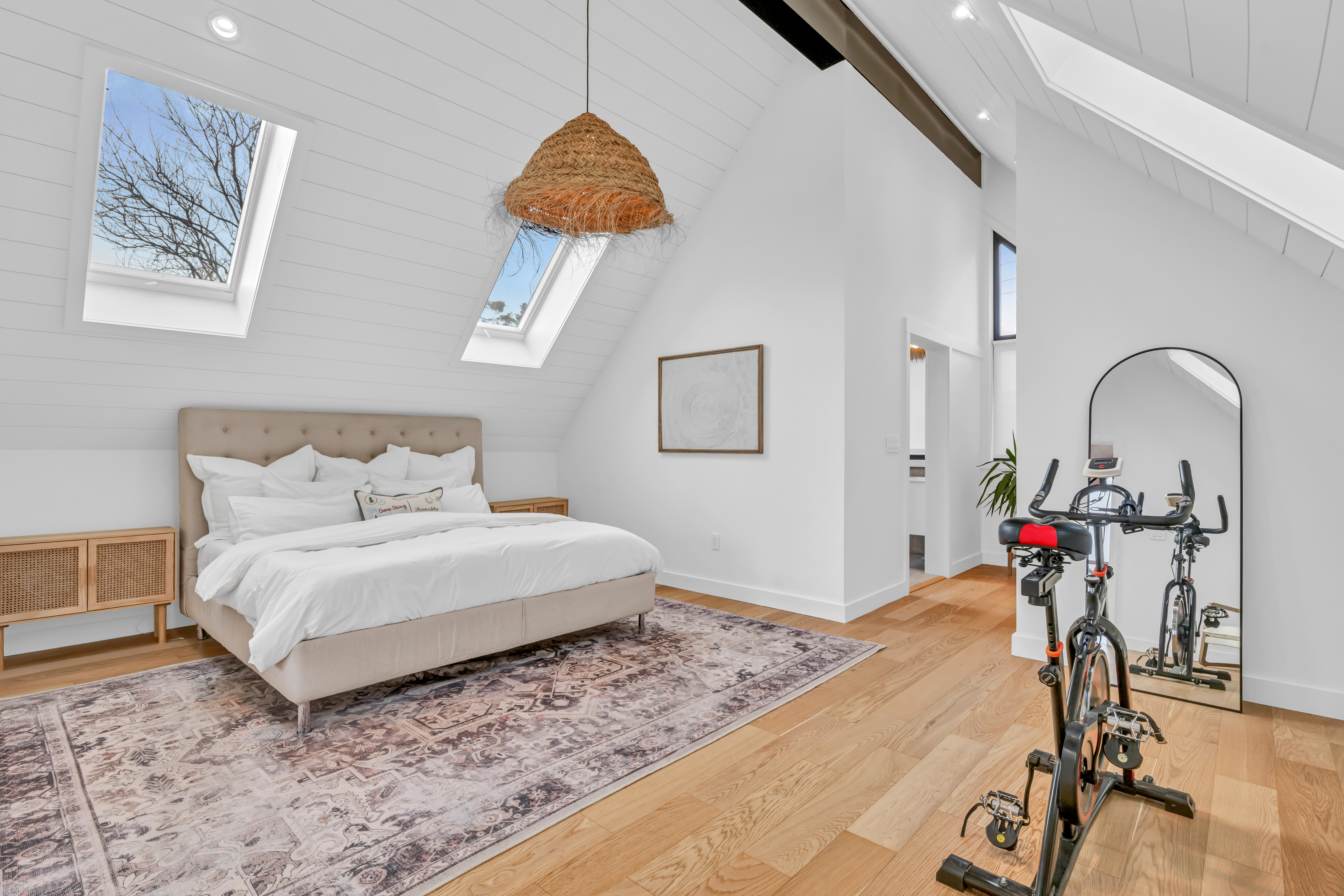 A big bed in a bright, open room, on top of a large area rug on a hardwood floor. The slanted ceiling has skylights, letting in lots of natural light. There is an exercise bike and a mirror across from the bed, and a short, narrow hallway leads to an ensuite bathroom.