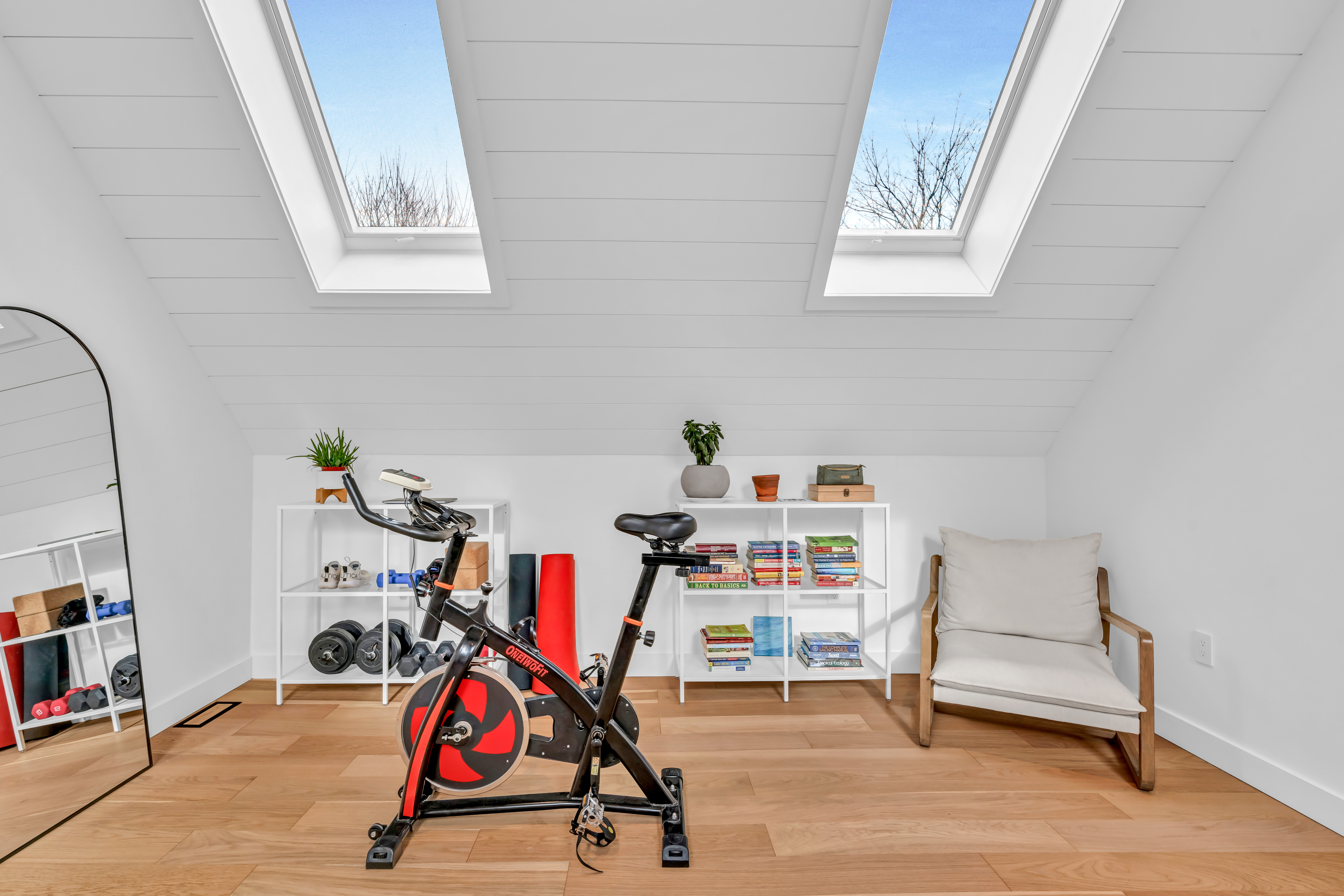 A bright space with a mirror, an exercise bike, short bookshelves and a chair. The slanted ceiling has two skylights, letting in lots of natural light.