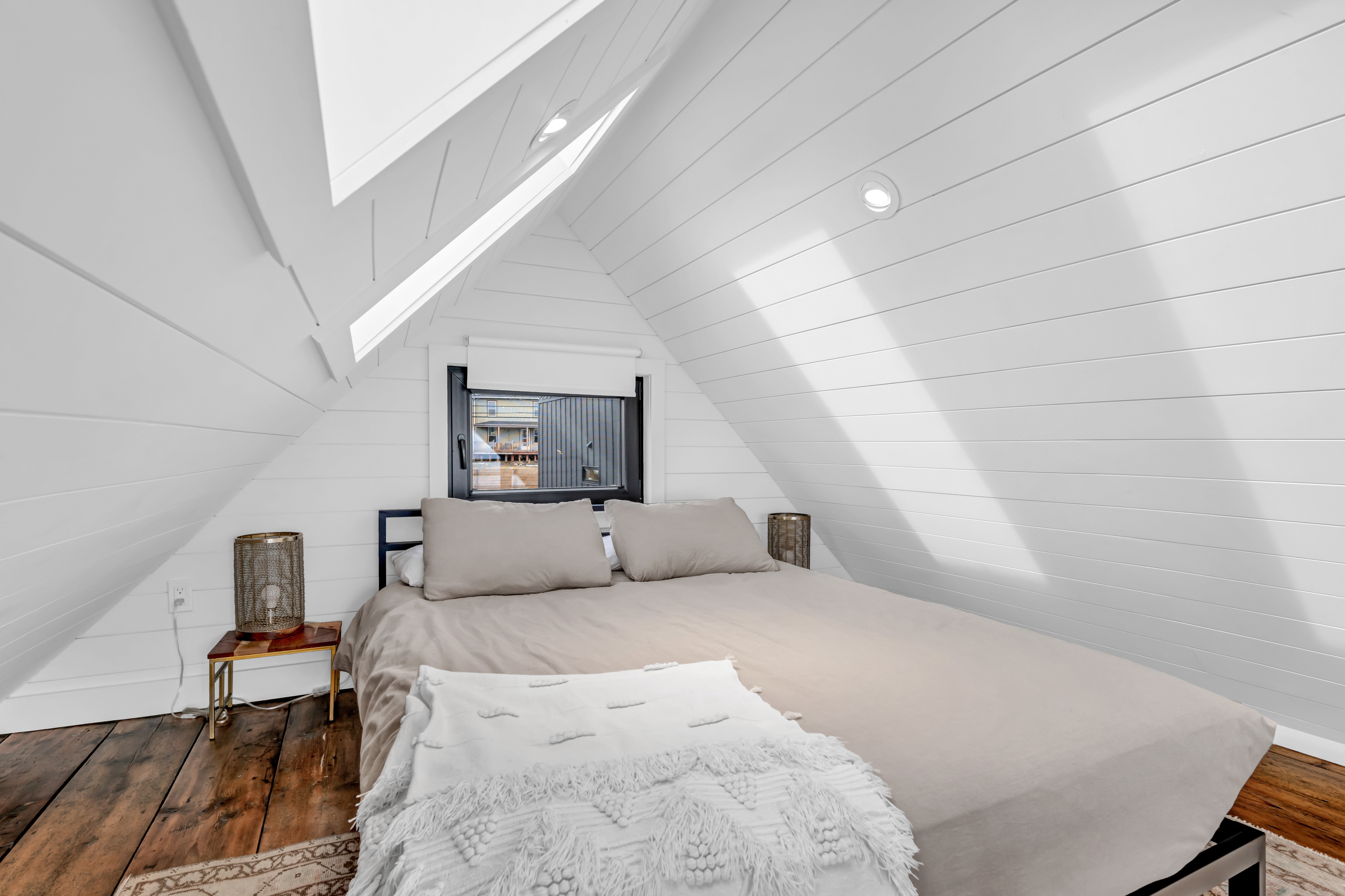 A bright loft bedroom area with a big bed, hardwood flooring, and a white-painted shiplap ceiling with skylights.