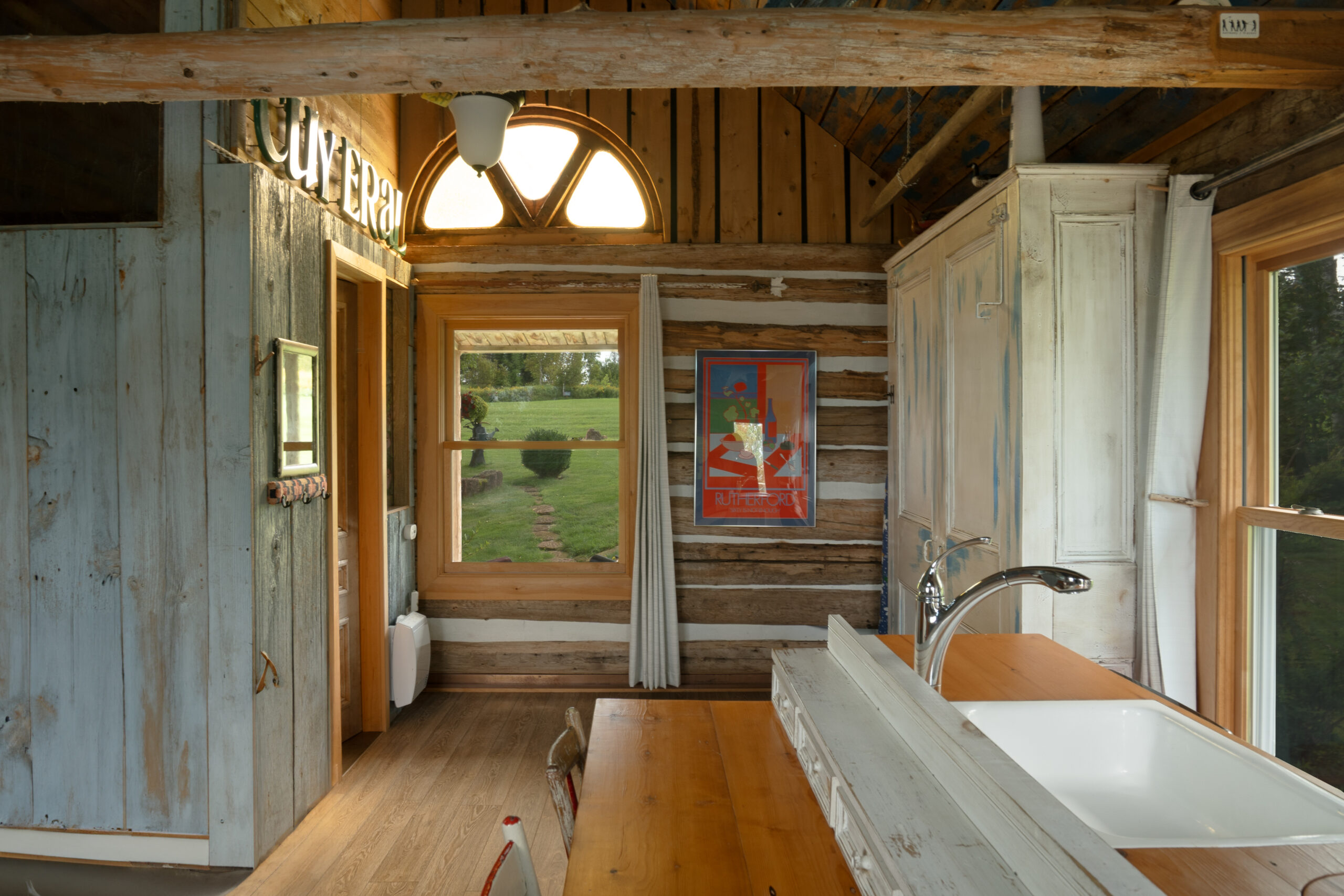 Interior of a small log cabin, with a table, a big window, a sink, and a storage cupboard.