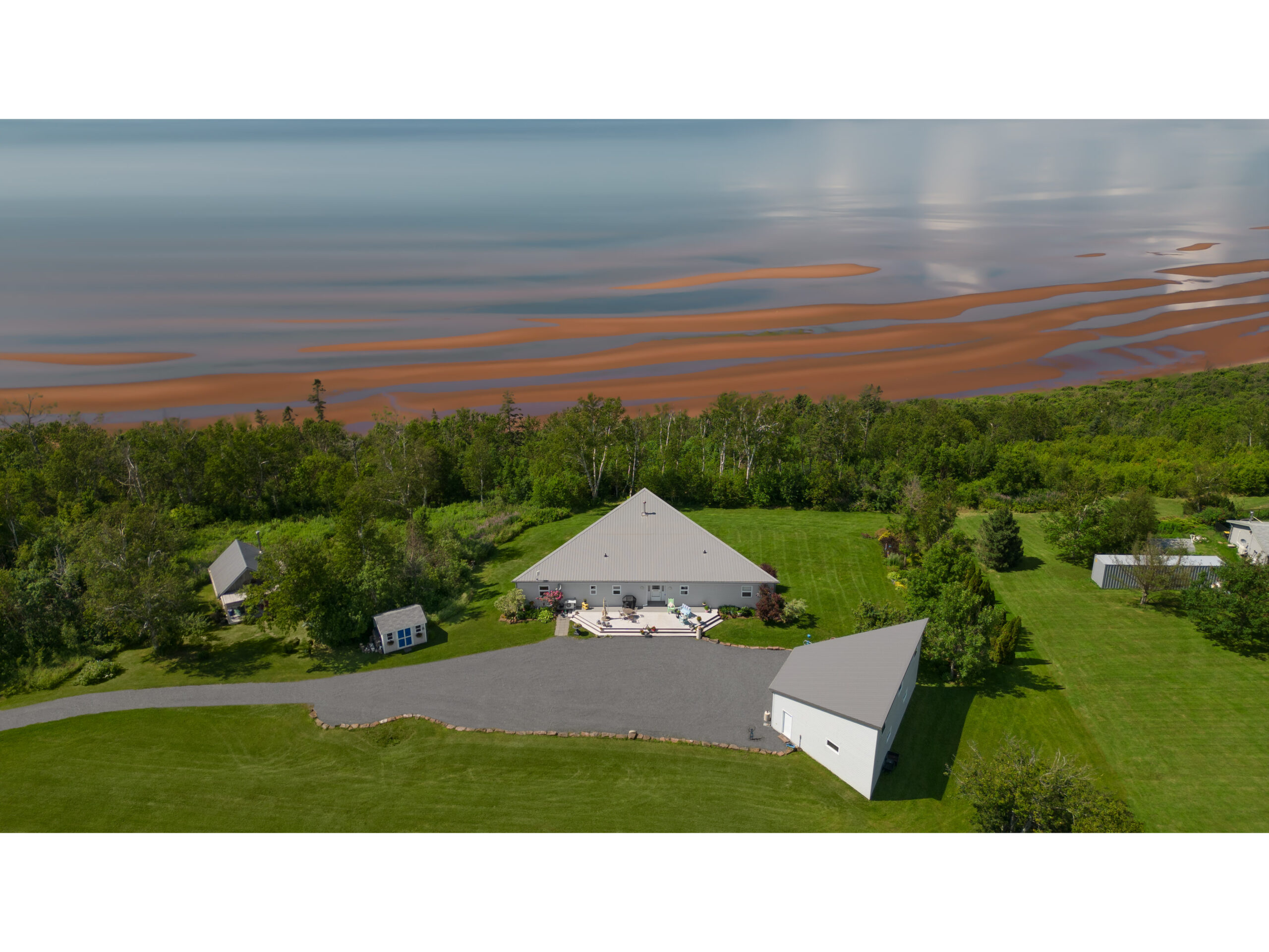 A large waterfront Prince Edward Island home surrounded by green trees, looking out to a red sand beach and a big stretch of water.