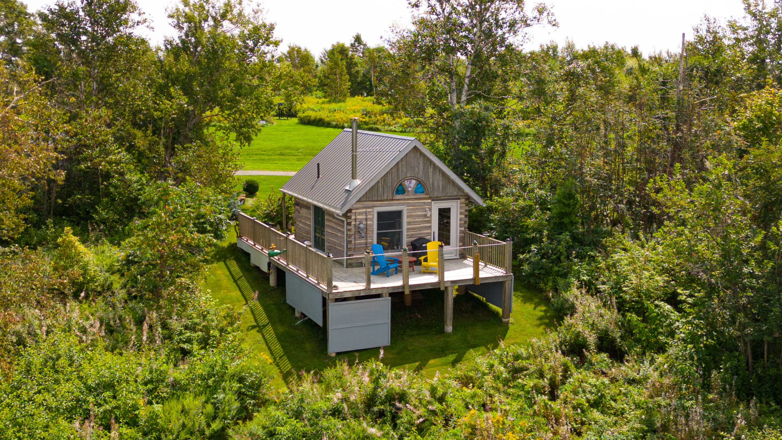 A small log cabin with a modern wrap-around deck, which holds two Muskoka-style chairs.