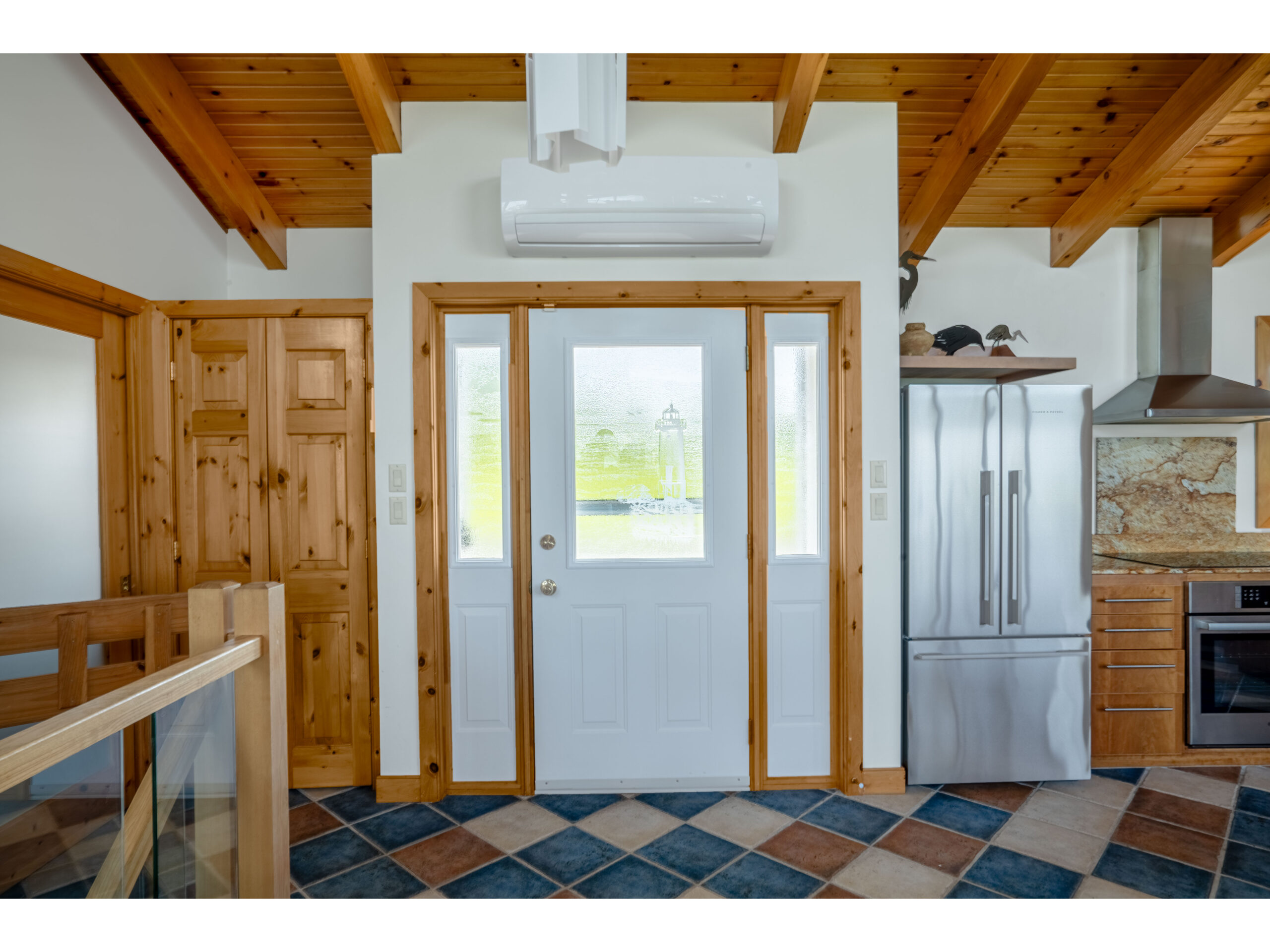 A big front entrance way with a white door framed in wooden trim. To the right is a fridge in the kitchen. The floor is tiled with red, blue, and beige tiles.