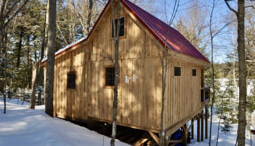 A one and a half story timber frame cabin with a red roof sits in a snowy wooded area.
