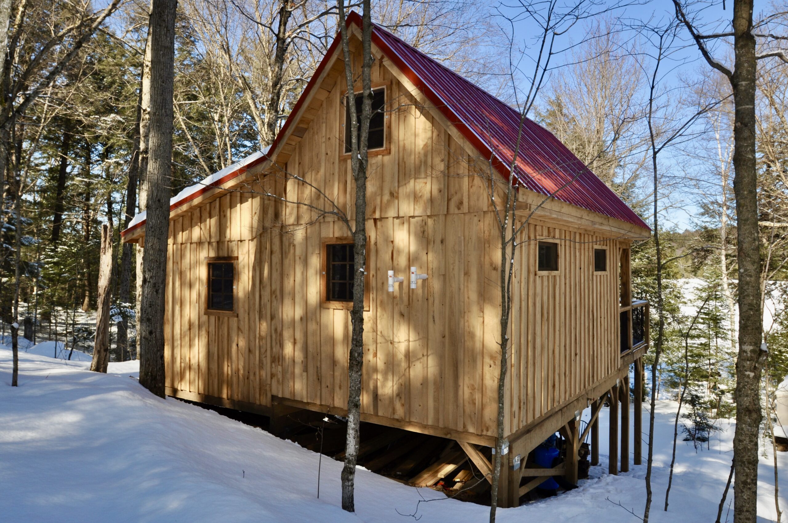 A one and a half story timber frame cabin with a red roof sits in a snowy wooded area.