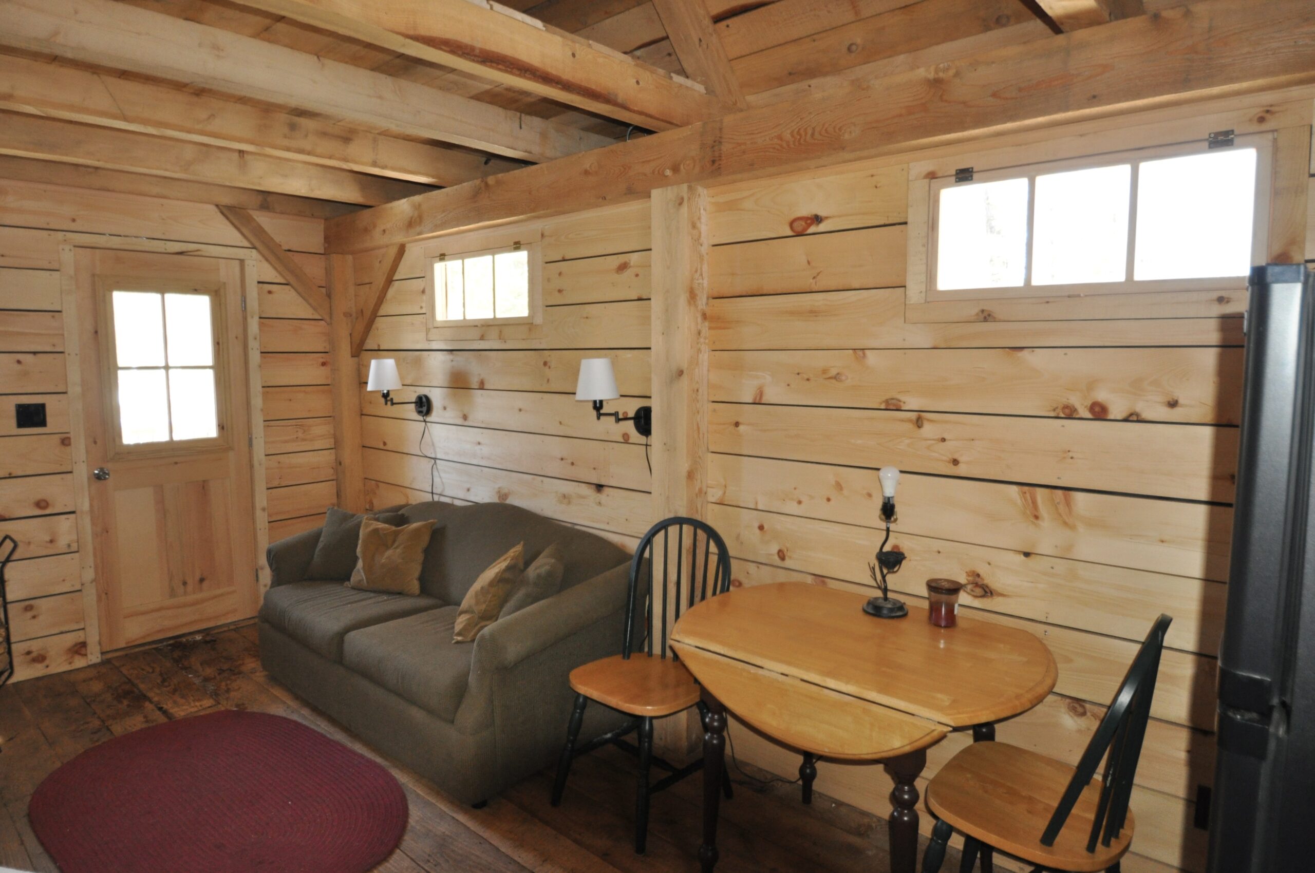 Interior living area of a wood cabin, with a couch and a small dining table. Small windows let in natural light.