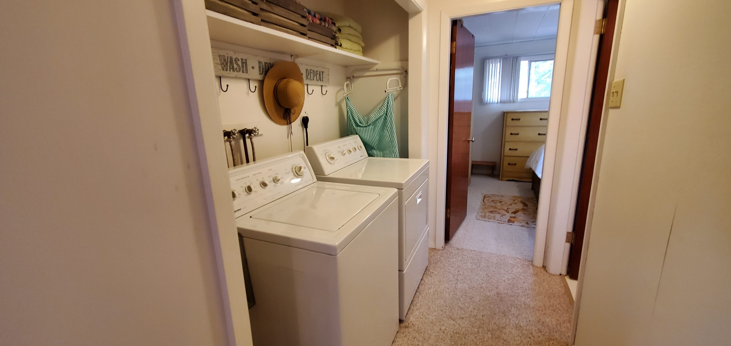A washing machine and a dryer sit side by side in a hallway alcove. Hooks hang on the wall above them, with a shelf above the hooks.