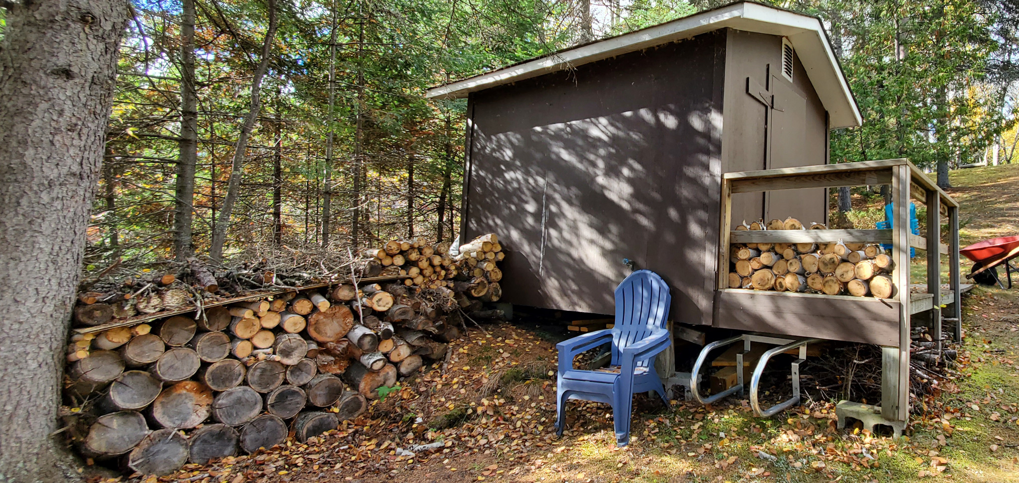 A small storage shed. A pile of fire wood and a blue Muskoka chair sit beside the shed.