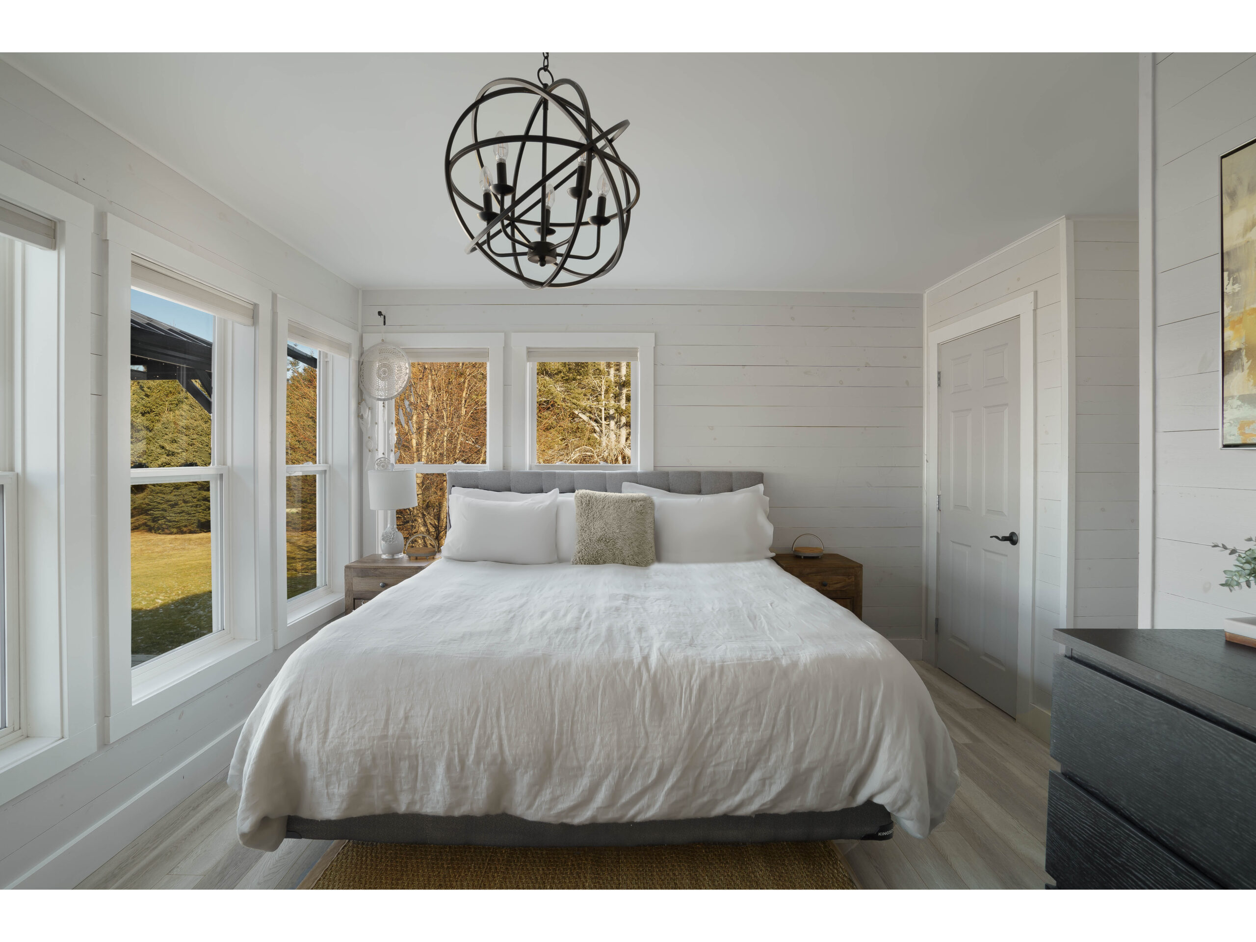 A large primary bedroom with a big bed, a modern light fixture, a dresser, and lots of windows.
