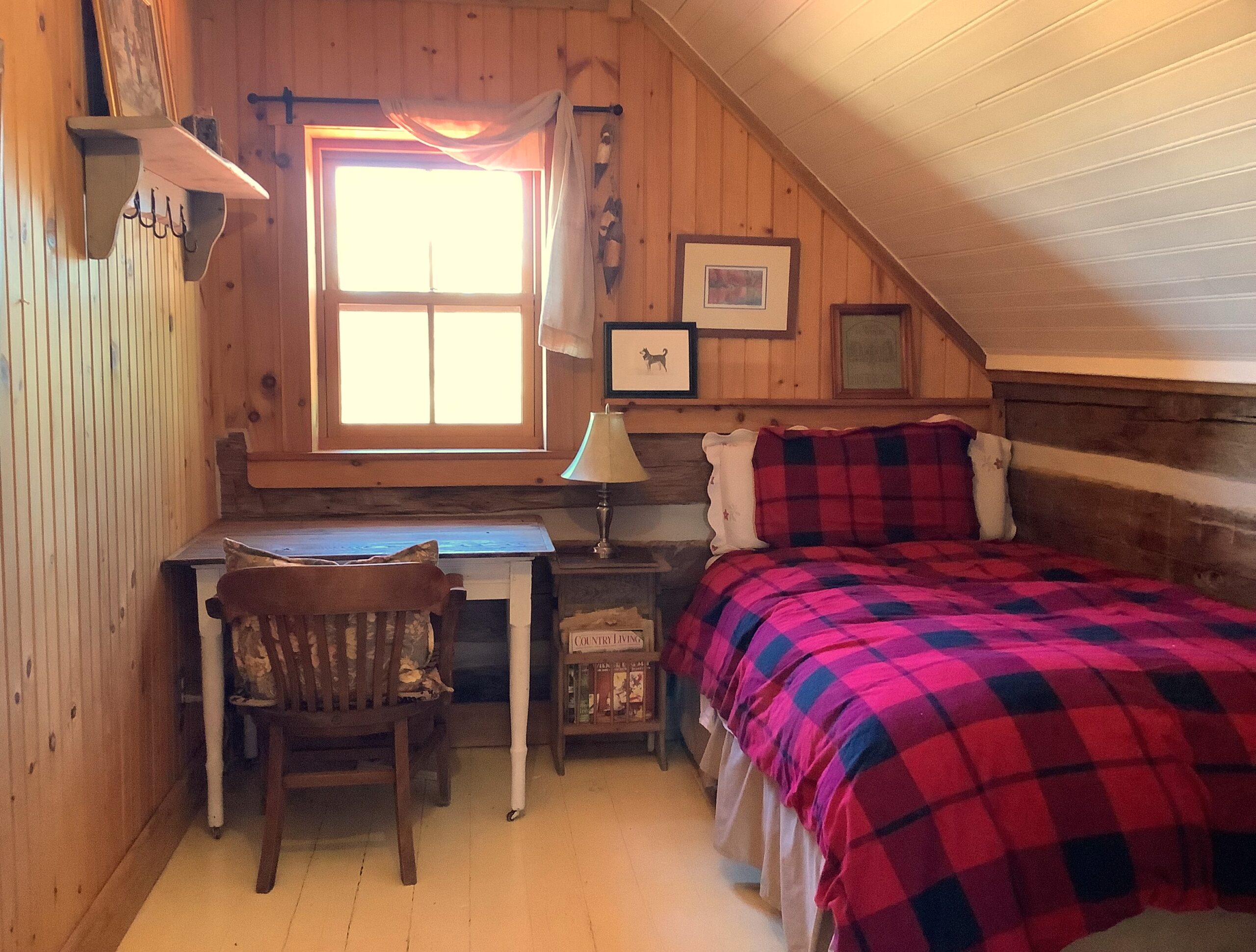 A small bedroom with one wall sloping toward the ceiling. The bedroom has a window, a small desk, and a bed with a red-and-white flannel patterned bedspread.