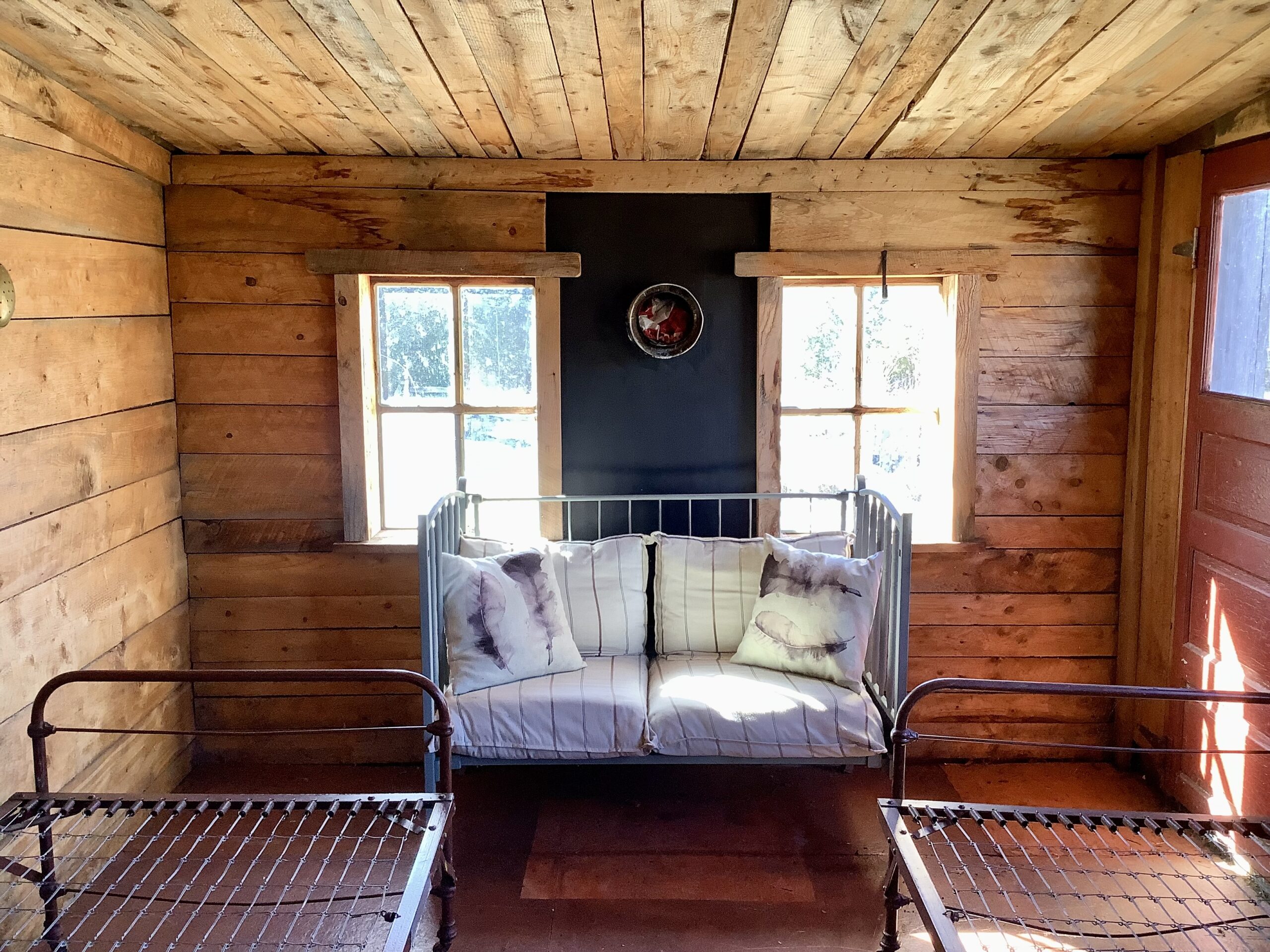 Interior of a small, rustic wooden bunkie with two empty metal bed frames, two windows, and a small couch.