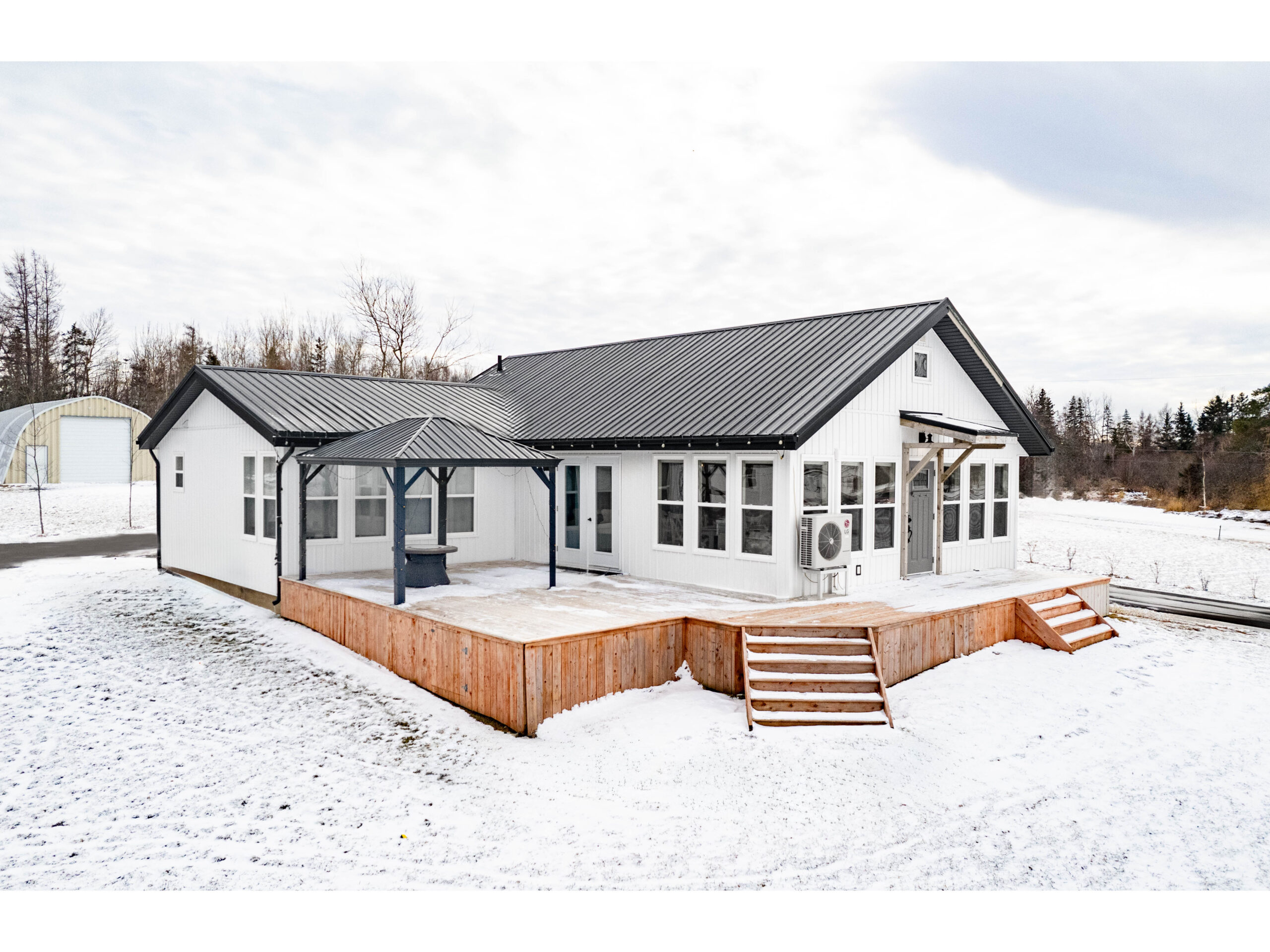 A small home with a white exterior, black trim, a big wooden deck, and lots of windows, sitting on a snow-covered lot.