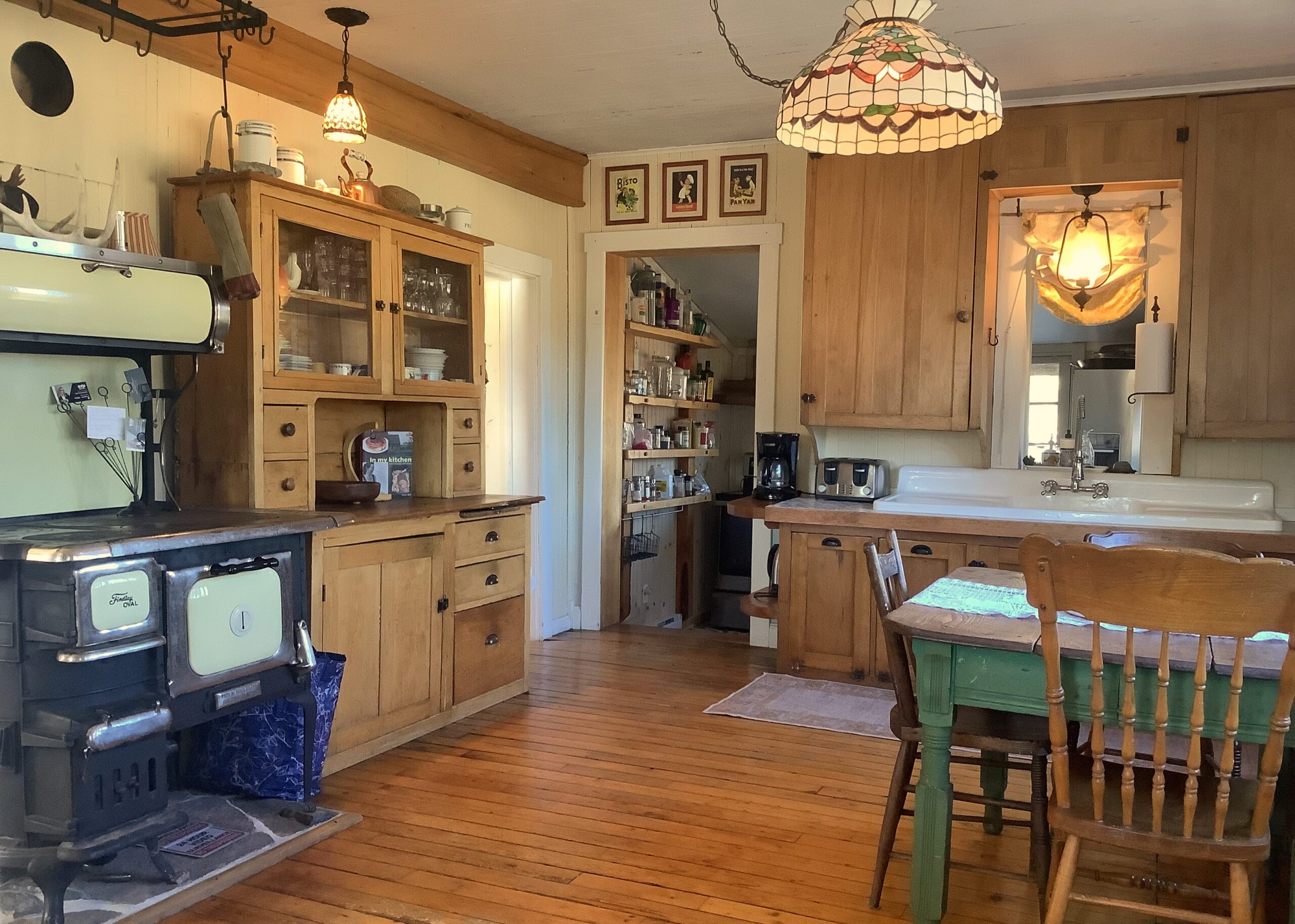 A kitchen with hardwood flooring, a small table and chairs, wooden cabinets, a vintage glass light fixture, and an old stove.
