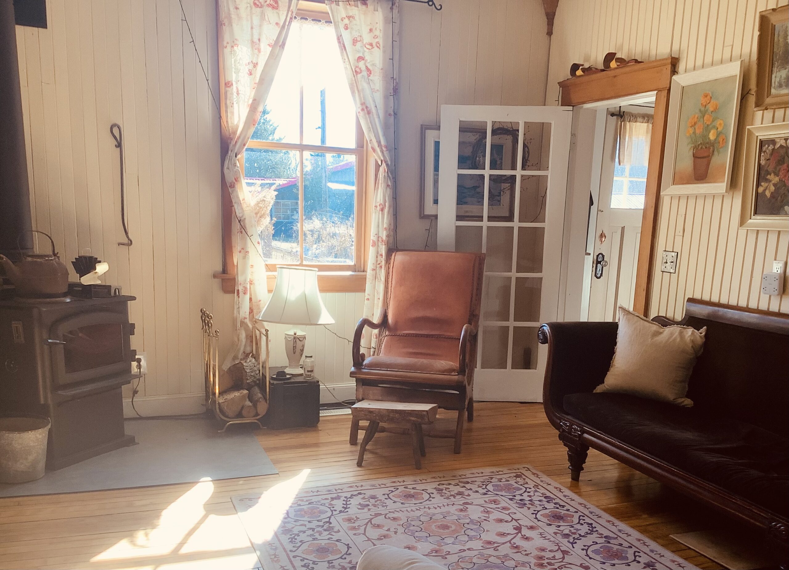 A small, bright living area with a woodstove in the corner, a window with drapes pulled to either side, a pink chair, a vintage couch, and an ornate floral rug over hardwood flooring. A gallery wall of framed art hangs above the couch.