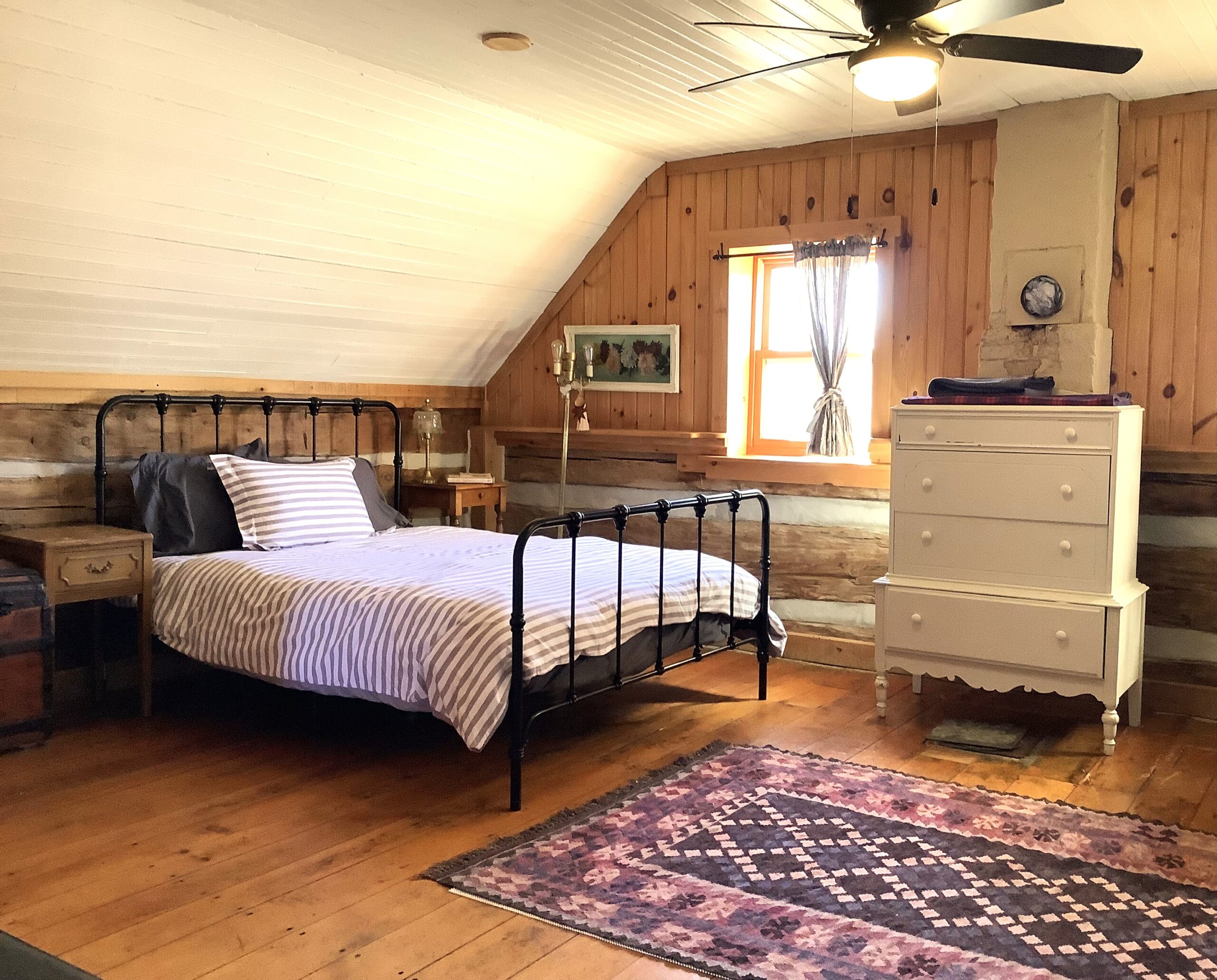 A big bedroom with angled walls, a big bed, a white dresser, a ceiling fan, and an ornate rug over hardwood flooring.