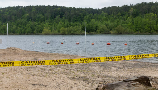 Caution tape restricts beach access in front of a lake.